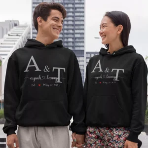 Couple wearing black personalised hoodies with A & T initials, custom names and date, holding hands outdoors.